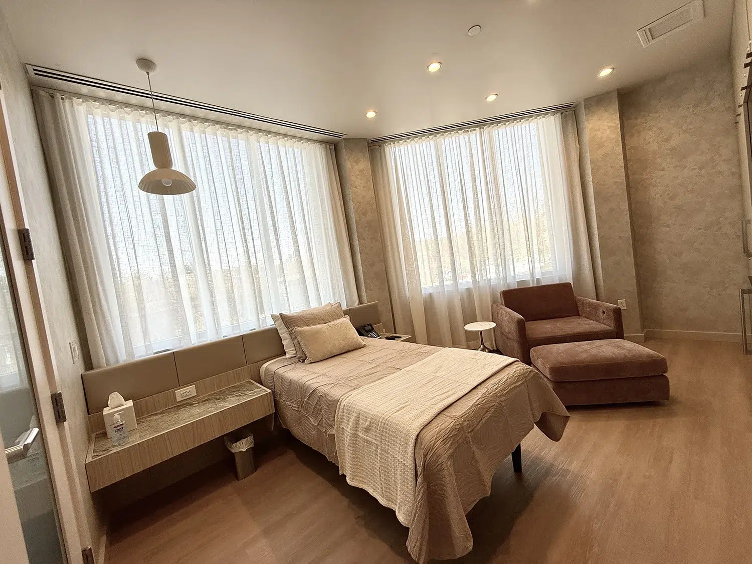 Spacious medical recovery room with large windows, sheer curtains, adjustable bed, and modern pendant lighting.